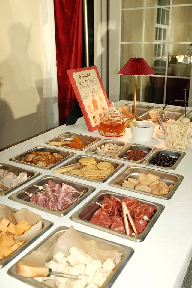 The charcuterie cart's steel pans of crackers, salami, cheese and olives, with a honey jar and a red lamp