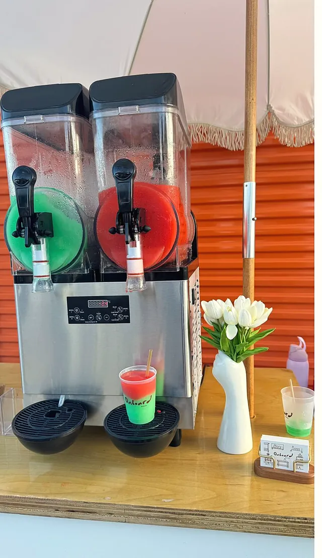 The slush machine with red and green flavours, flowers and a cup, against an orange wall